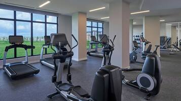 Une salle de sport équipée de tapis de course, de vélos elliptiques et offrant une vue sur un champ verdoyant grâce à de grandes fenêtres.