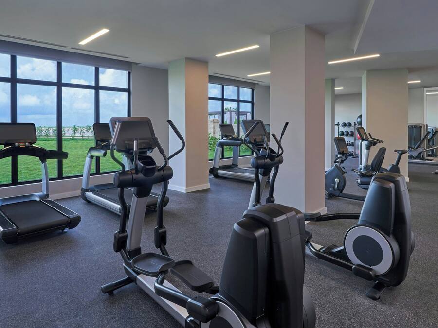 A gym with treadmills, elliptical machines, and a view of a green field through large windows.