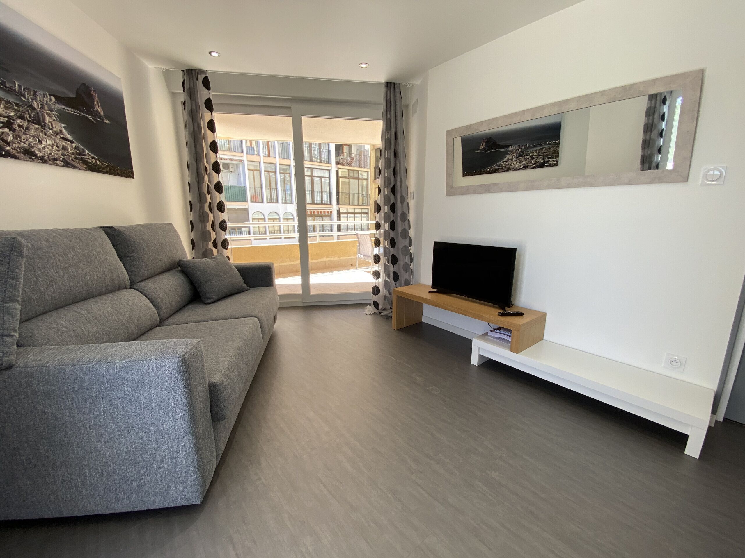 A modern living room with a grey sofa, a TV on a wooden stand, and a balcony with a view of buildings.