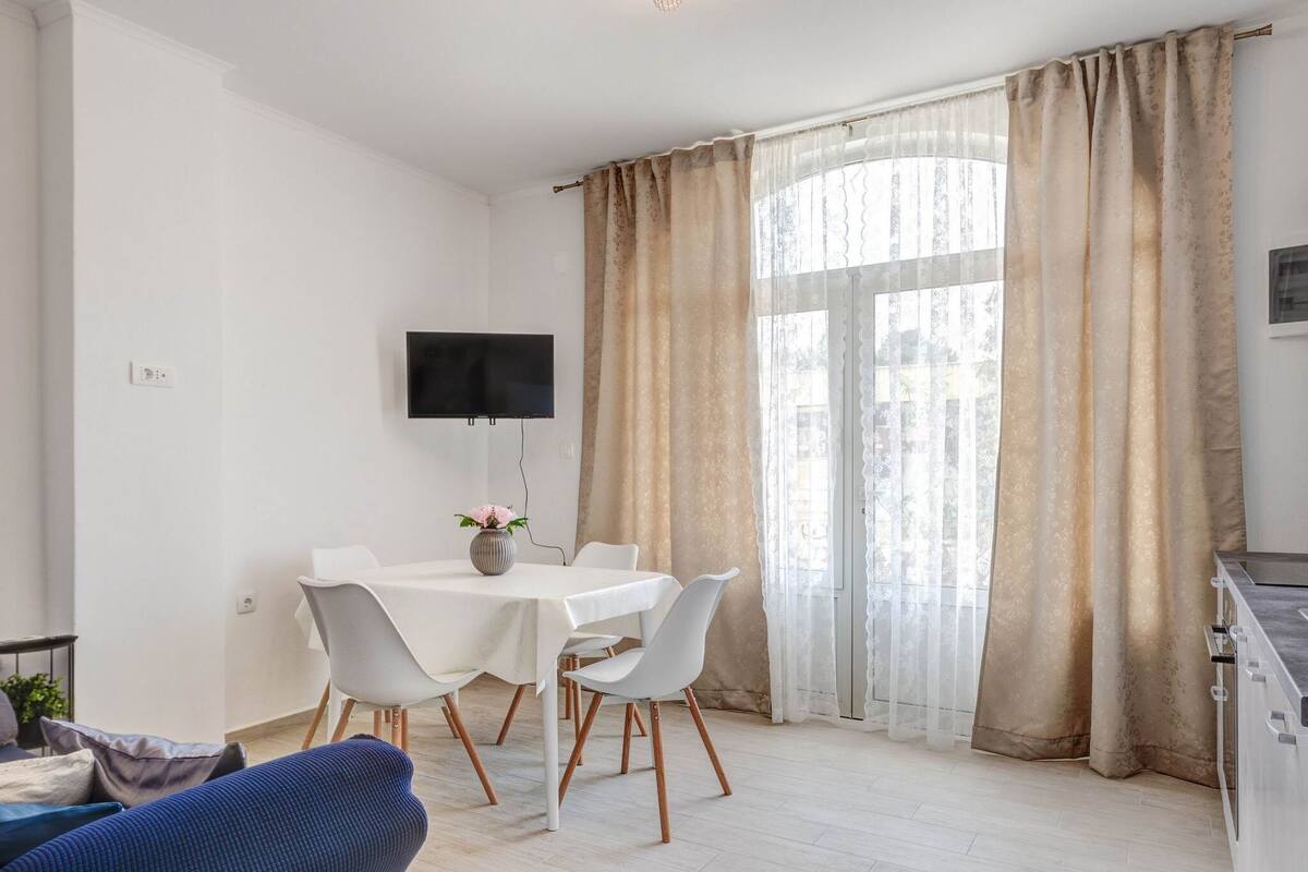 A modern, well-lit room with a dining table, chairs, a TV mounted on the wall, and large windows with curtains.