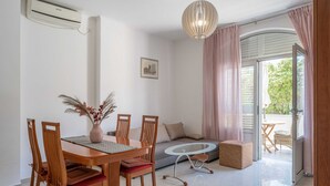 A dining area with a wooden table and chairs, a glass-top coffee table, and a sofa. There is a window with curtains, a door leading to an outdoor area, and an air conditioning unit on the wall. - Apartments K & N (Blato)
