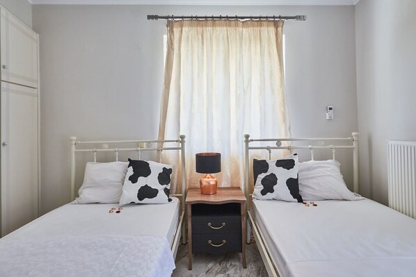 Un dormitorio con dos camas individuales, una mesita de noche con lámpara y una ventana con cortinas.