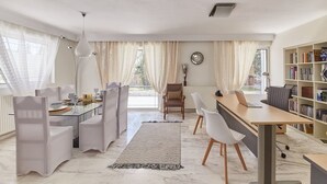 A modern living room with a dining area, a bookshelf, and a large window with curtains. - Harmony Vale- Anavissos (Saronikos)