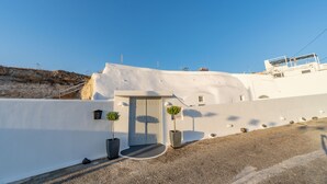 A white building with a door and two potted plants, a gravel path, and a clear blue sky. - Fira-cave-sleeps 13-parking-pool-pet Friendly (Santorini)