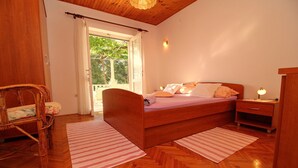 A bedroom with a wooden bed, a nightstand, a chair, and a window with curtains. - Apartment Kim (Lumbarda)