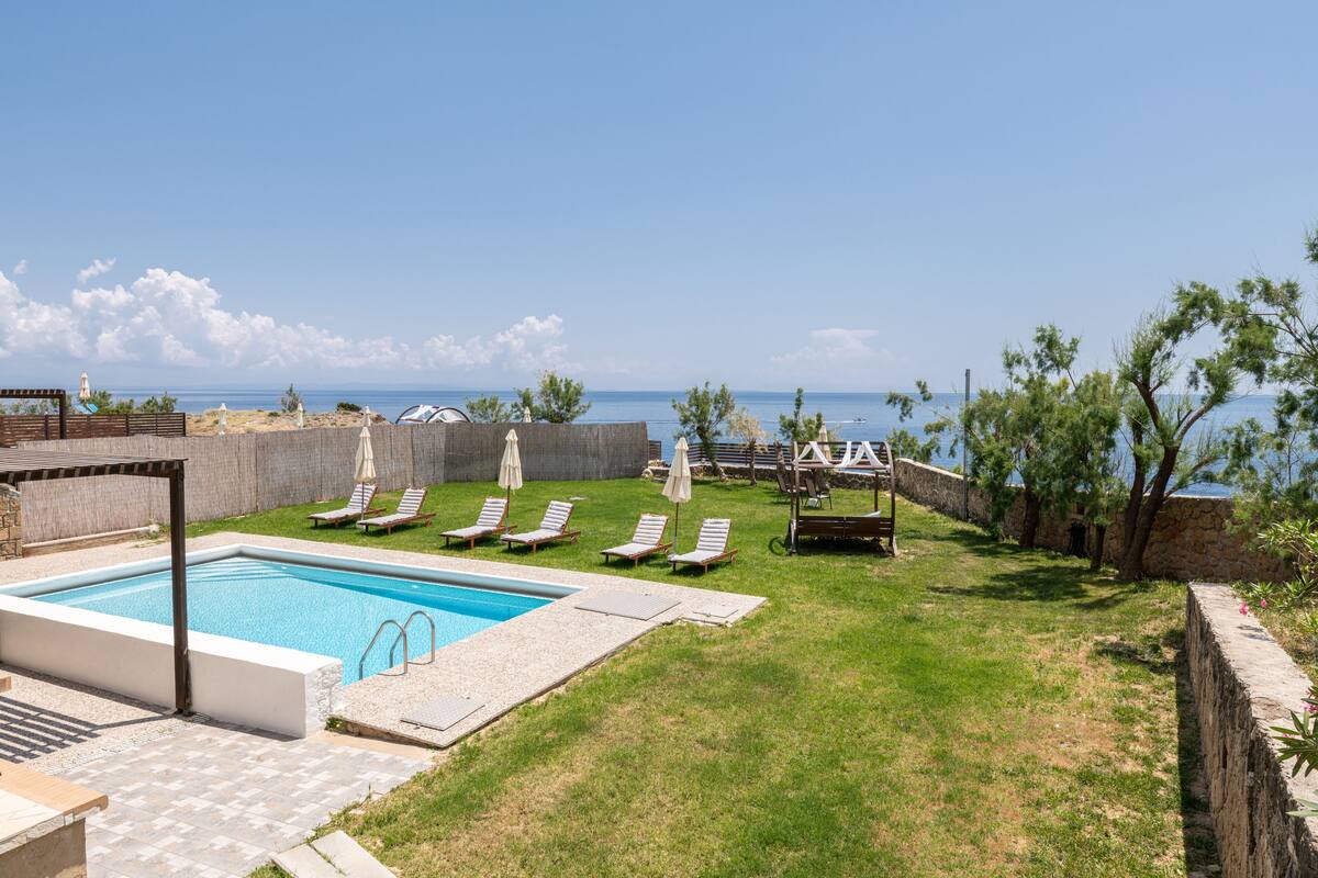 A swimming pool with lounge chairs and umbrellas, a stone wall, and a view of the sea.