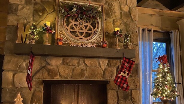 A stone fireplace with a fire, decorated with Christmas ornaments and stockings. - Sweets River Nest-riverfront/tube/hot tub/log cabin-fishing- Valle Crucis/App ST (Banner Elk)