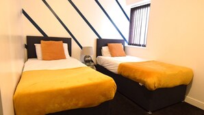 Ein Hotelzimmer mit zwei Betten, jedes mit einem schwarzen Kopfteil und einer gelben Tagesdecke. Dazwischen steht ein kleiner Tisch mit einer Lampe.
