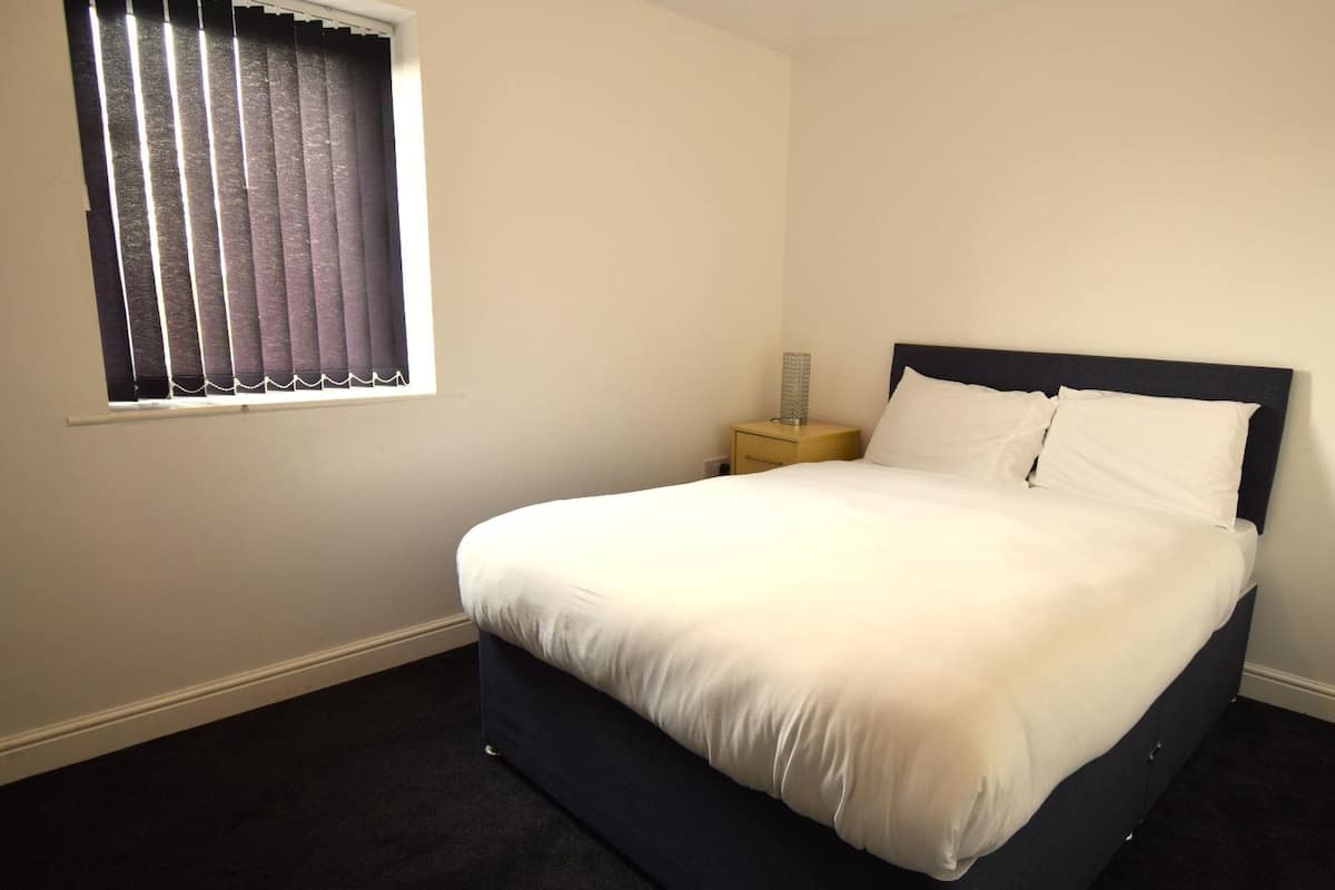 A bedroom with a bed, a nightstand, a lamp, and a window with blinds.