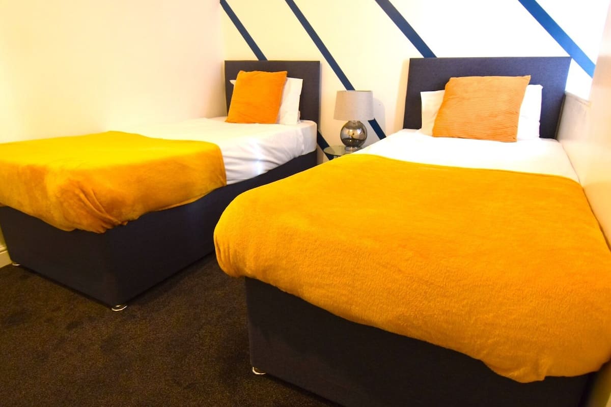 Two single beds with orange bedding, a nightstand with a lamp, and a wall with a blue and white geometric pattern.