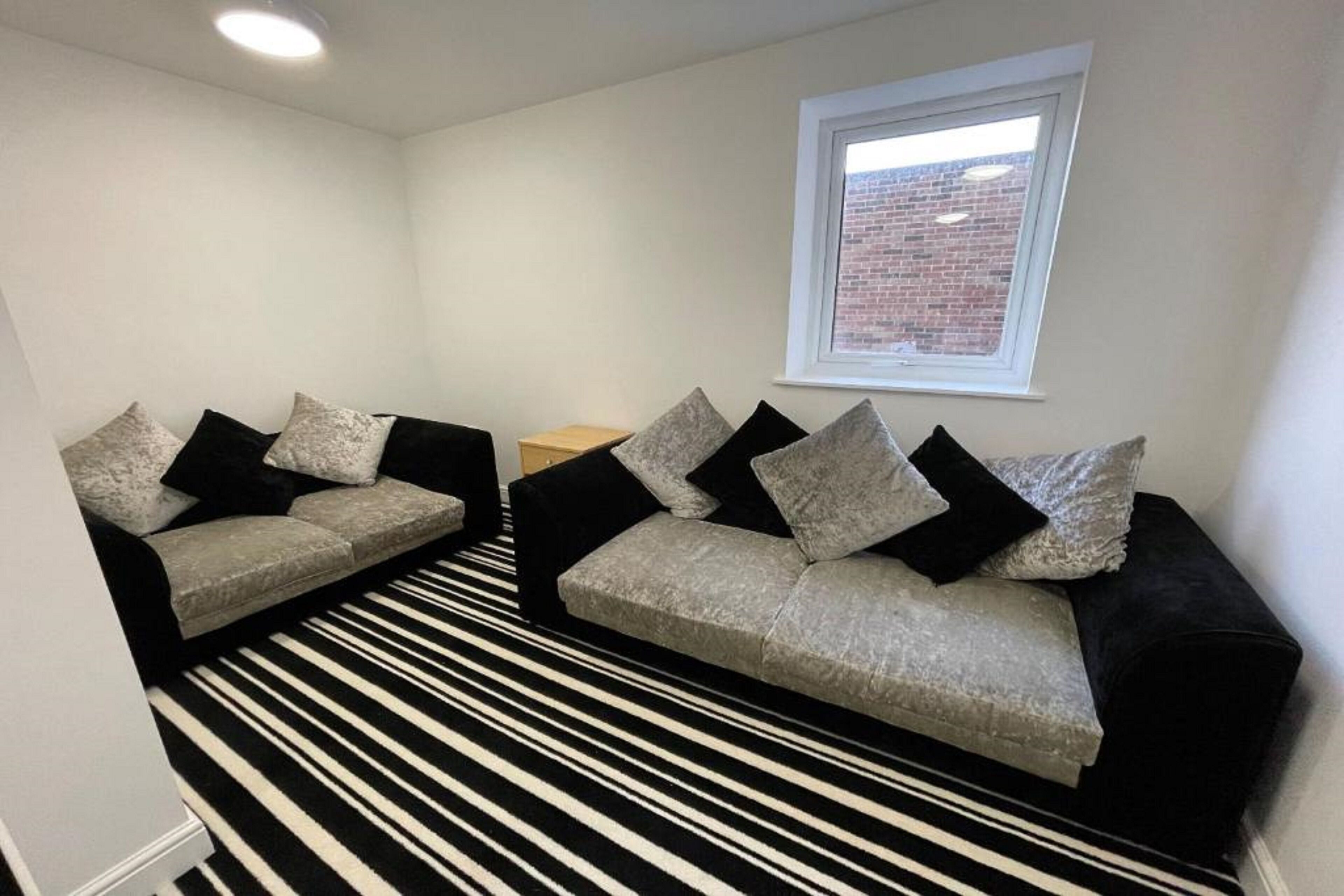 A room with two grey sofas, black and white striped carpet, a small wooden table, and a window with a brick wall view.