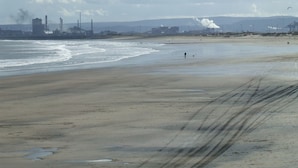 Ein weiter Sandstrand mit einer in der Ferne liegenden Industrieanlage, einschließlich Schornsteinen und einem Kran.