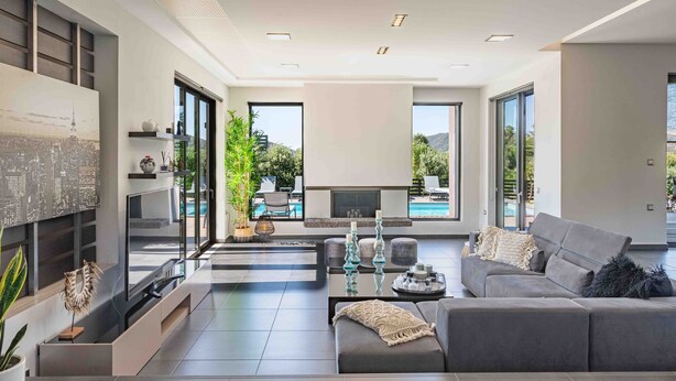 Un soggiorno moderno con un divano grigio, un tavolino da caffè e una televisione. Si nota un camino, una libreria e un'ampia finestra con vista su una piscina.
