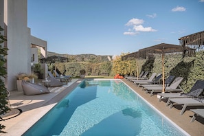 A swimming pool with lounge chairs and umbrellas, surrounded by greenery and hills in the background. - Villa Vega (Platanias)