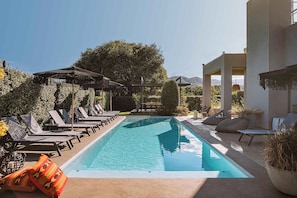 A swimming pool with lounge chairs and umbrellas, surrounded by greenery and a mountainous backdrop. - Villa Vega (Platanias)