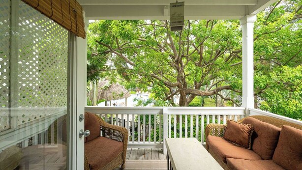 A covered outdoor seating area with wicker furniture and a white table, overlooking a large tree and lush greenery.