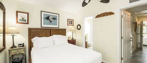 A bedroom with a large bed, a ceiling fan, framed pictures on the wall, and a door leading to another room.