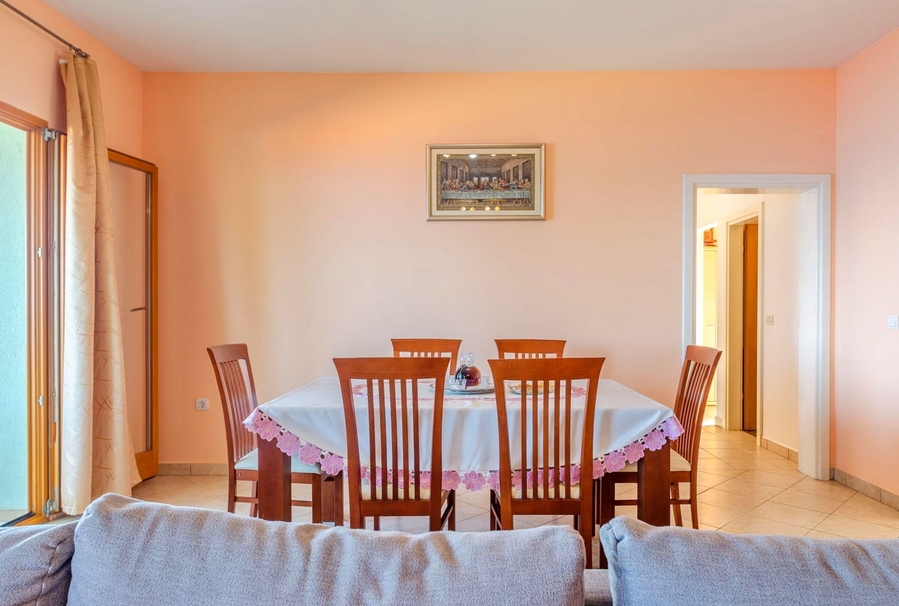 A dining area with a table set for four, surrounded by wooden chairs, a framed artwork above the table, and a view of a hallway.