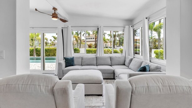 A modern living room with a grey sectional sofa, a ceiling fan, and large windows offering a view of a pool and greenery. - The Palm Leaf 3 Bedroom Home by RedAwning (Anna Maria)