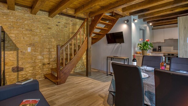 Un intérieur rustique avec des poutres apparentes, un mur en pierre, un escalier en bois, un coin repas avec une table et des chaises, et un coin cuisine avec une télévision et une plante.