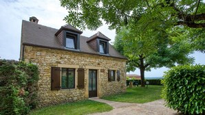 Une maison en pierre avec une allée gravillonnée, un petit jardin et un arbre au premier plan.