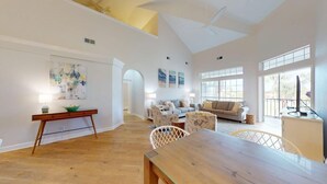A spacious living area with a wooden dining table, a sofa set, and a large window providing natural light.