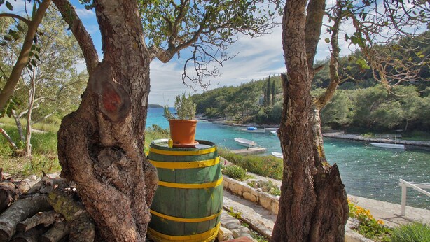 A rustic outdoor setting with a large barrel, a potted plant, and a view of a calm body of water with boats. - Apartment Slatina (Vela Luka)