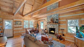 A spacious living room with a stone fireplace, wooden beams, and large windows.