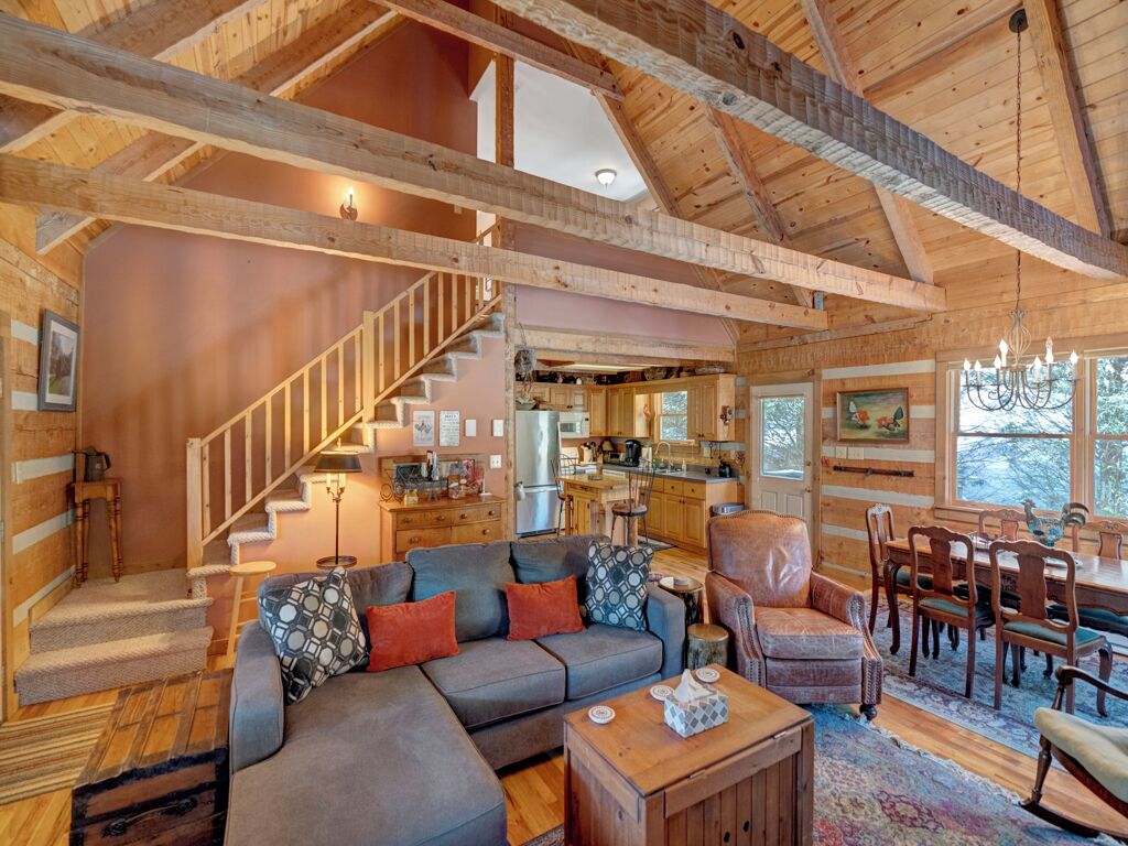 A spacious living area with a wooden ceiling, a sofa set, a dining table with chairs, and a kitchen in the background.