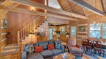 A spacious living area with a wooden ceiling, a sofa set, a dining table with chairs, and a kitchen in the background.