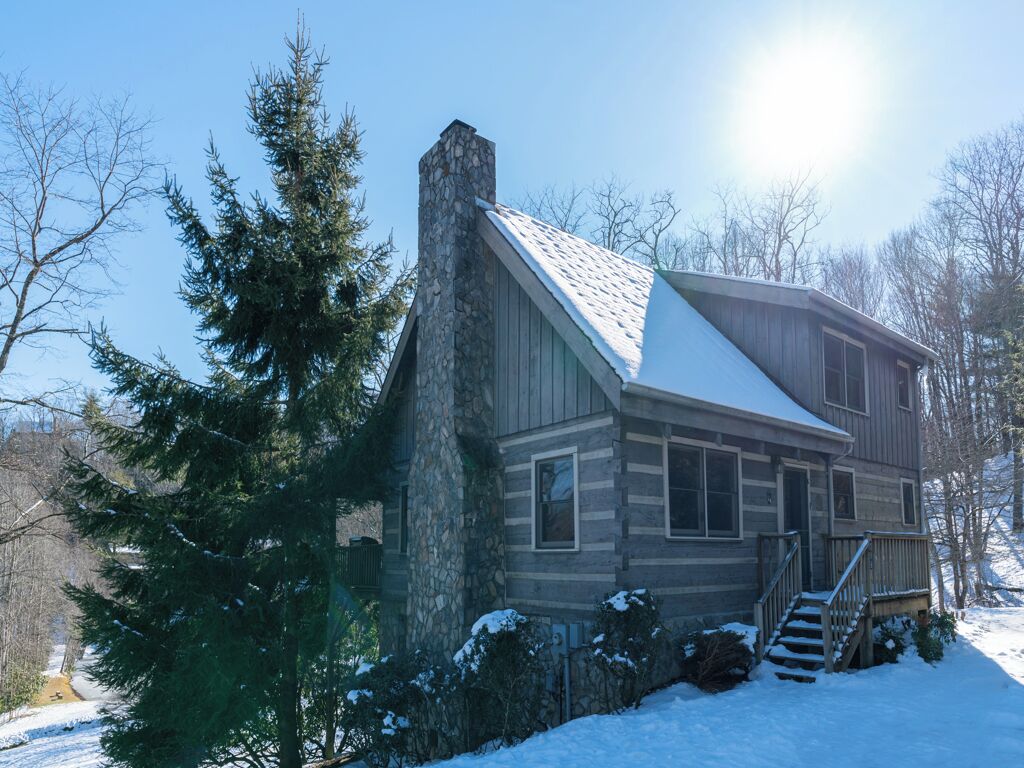 A log cabin with a stone chimney, a snow-covered roof, and a wooden deck.