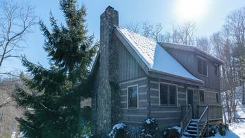 A log cabin with a stone chimney, a snow-covered roof, and a wooden deck.