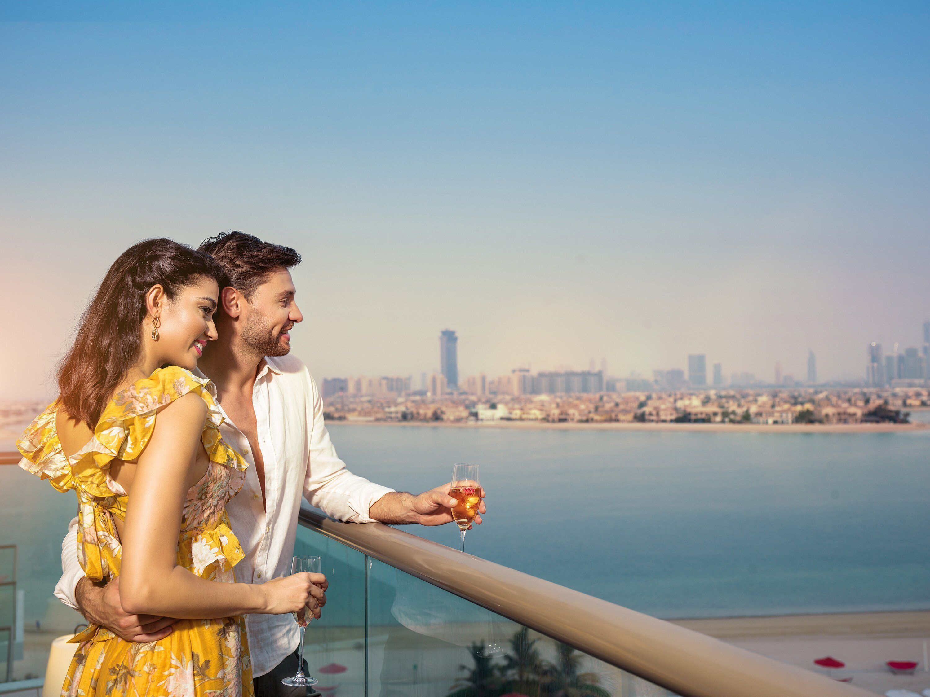 Una pareja abrazándose en un balcón con vistas a un paisaje urbano y una masa de agua al frente.
