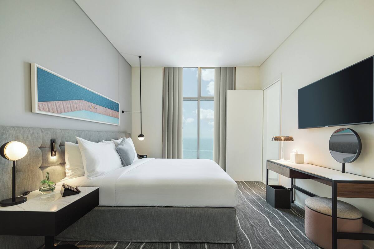 Une chambre d’hôtel moderne dotée d’un grand lit, de tables de chevet, d’un bureau, d’une télévision à écran plat et d’une grande fenêtre donnant sur un paysage agréable.