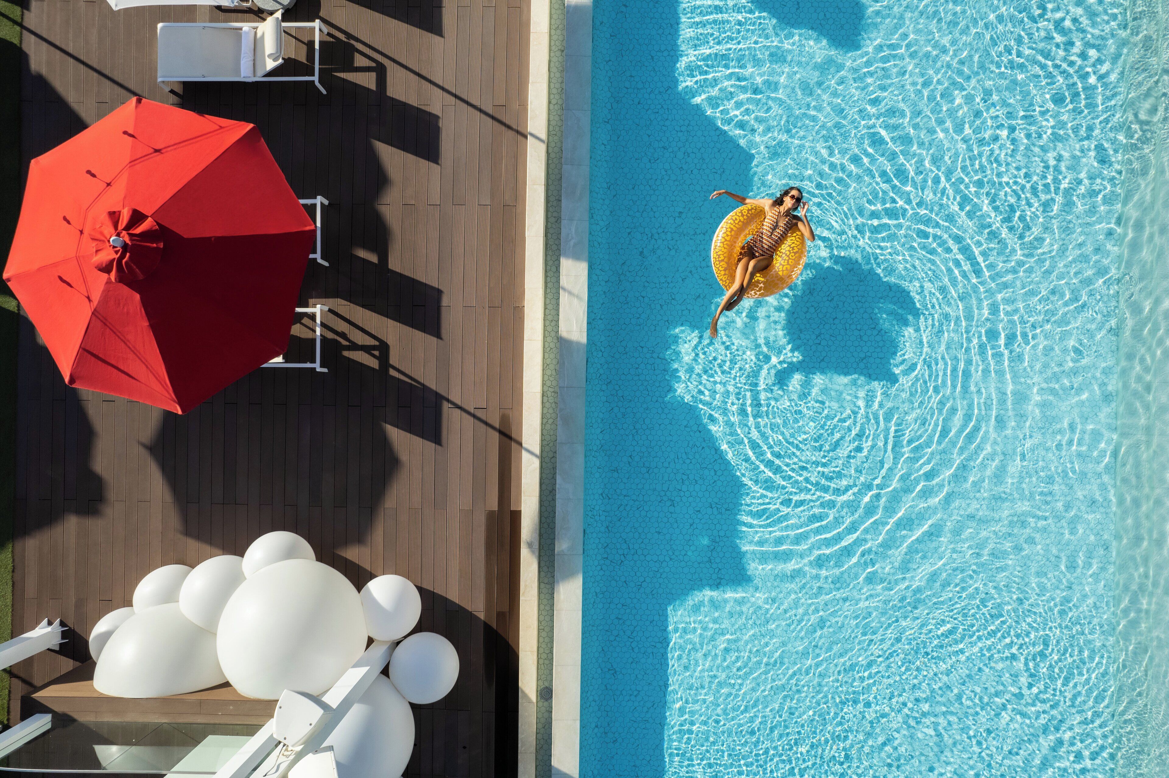 Vista aérea de una piscina con una persona en un flotador, un paraguas rojo y globos blancos.