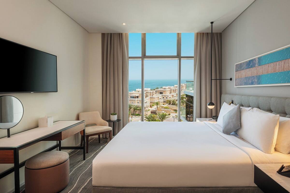 Une chambre d’hôtel avec un grand lit, un bureau, une chaise, une télévision et une vue sur la ville et la mer.
