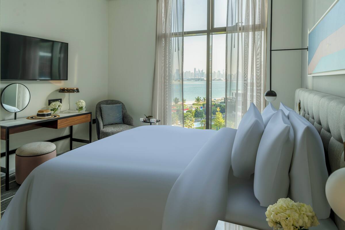 Une chambre d’hôtel moderne dotée d’un grand lit, d’un bureau, d’une chaise, d’une télévision et offrant une vue sur le paysage urbain et l’eau.