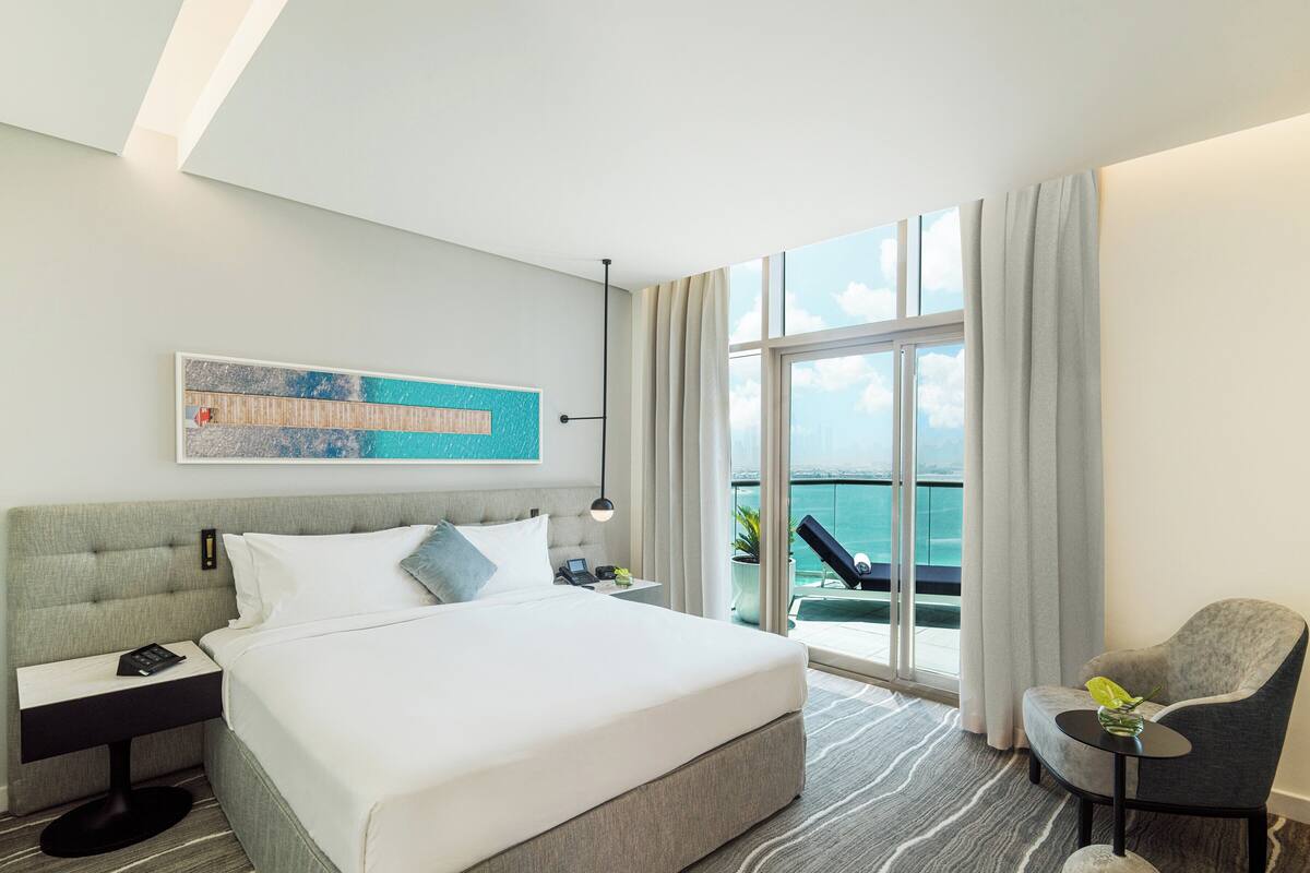 Une chambre d’hôtel moderne avec un grand lit, une table de chevet avec un téléphone, une chaise, une vue sur l’océan et un tableau au mur.