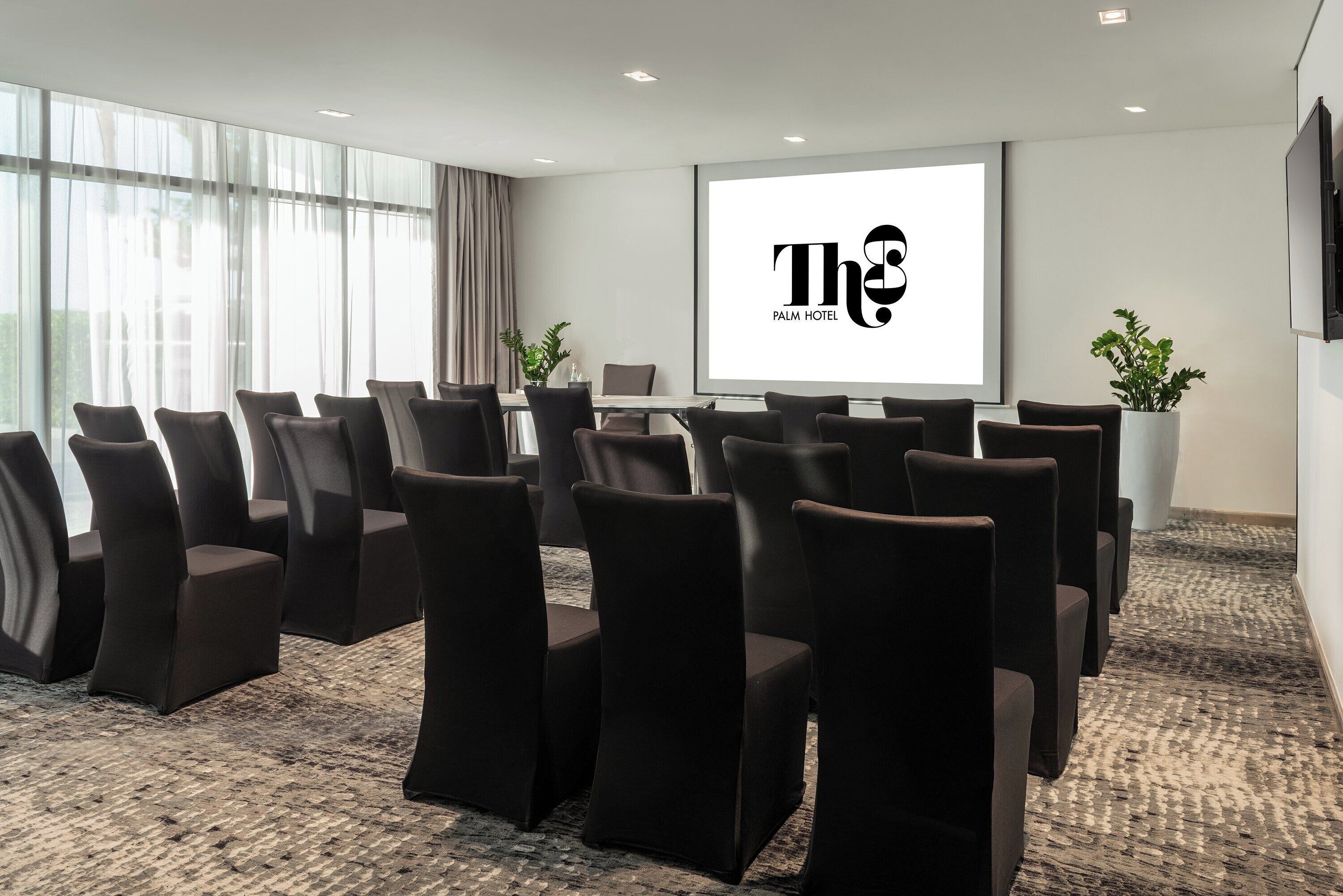 A conference room with a large screen displaying "The Palm Hotel" logo, black cushioned chairs arranged in rows, and a carpeted floor.