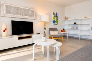 A modern living room with a flat-screen TV, a round coffee table, a grey armchair, a white lamp, and a wall-mounted shelf with decorative items. - Arenal Dream Penthouse I Javea Arenal  Luxury with Roof Terrace & only 150m from the Beach (Javea / Xàbia)