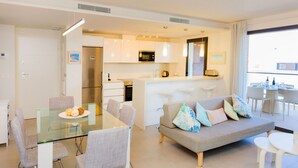 Een moderne keuken met witte kasten en een magnetron, een eethoek met een glazen tafel en stoelen, en een zithoek met een bank en kussens.