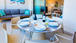 Een eetgedeelte met een ronde tafel voor zes personen, omringd door witte stoelen. Op de tafel staan borden, bestek en wijnglazen. Op de achtergrond is een grijze bank en een kleine tafel met een lamp te zien.