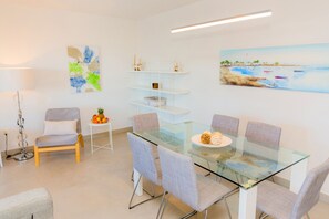 Een moderne eethoek met een glazen tafel, gestoffeerde grijze stoelen, een klein bijzettafeltje met fruit en een schilderij van een kustlandschap.