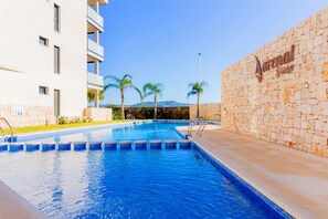 Piscina com muro de pedra e palmeiras, cercada por um edifício com varandas.