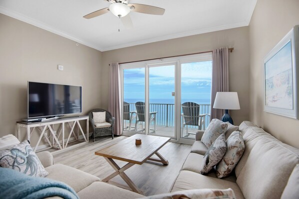 A living room with a flat-screen TV, a wooden coffee table, a sofa, and a balcony with a view of the ocean.