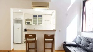 A compact living space with a kitchenette, two wooden chairs, a black sofa, and a window providing natural light. - Cool Duplex Studio Comfort & Great Location (São Paulo)