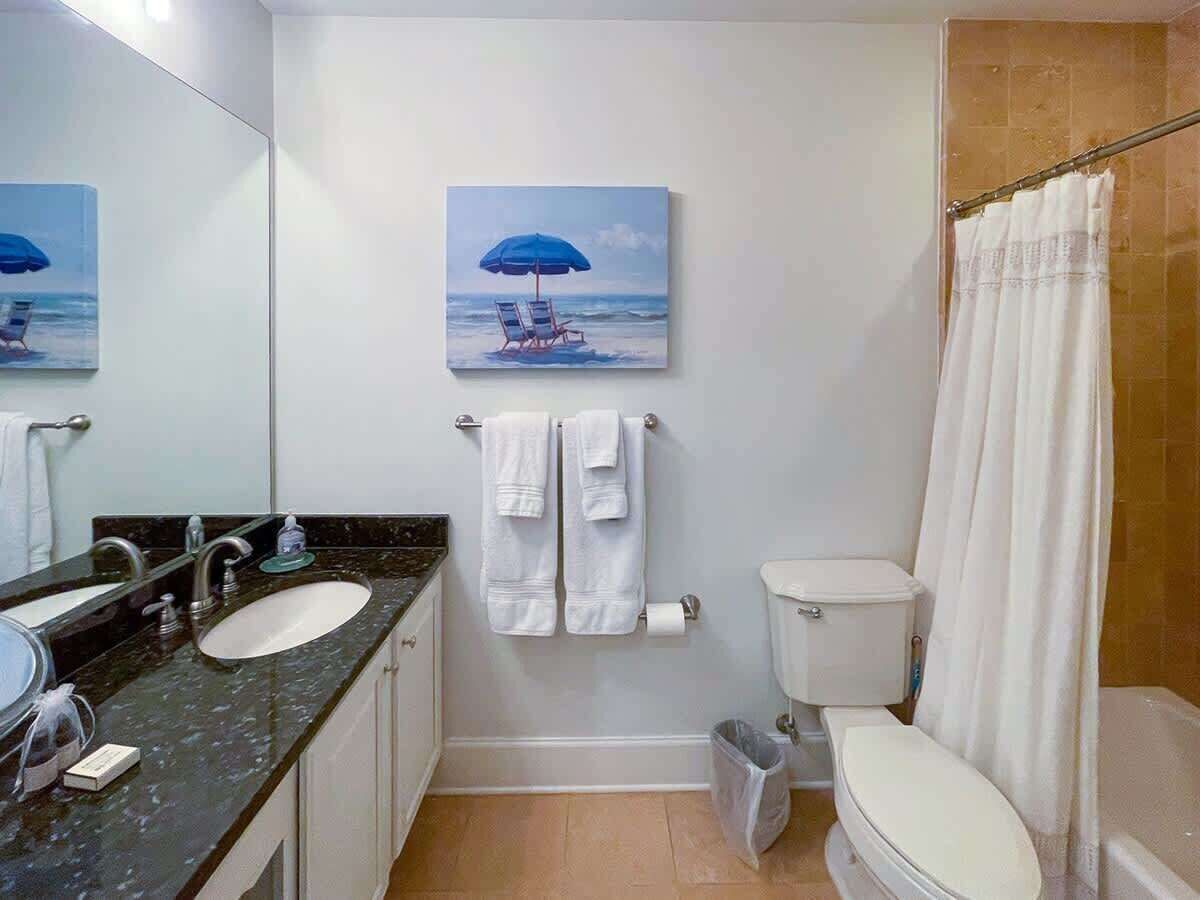 A bathroom with a sink, two towels, a toilet, a shower curtain, and a painting of a beach scene.