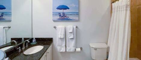 A bathroom with a sink, two towels, a toilet, a shower curtain, and a painting of a beach scene.