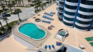An aerial view of a hotel with a large swimming pool, sun loungers, and a tennis court.