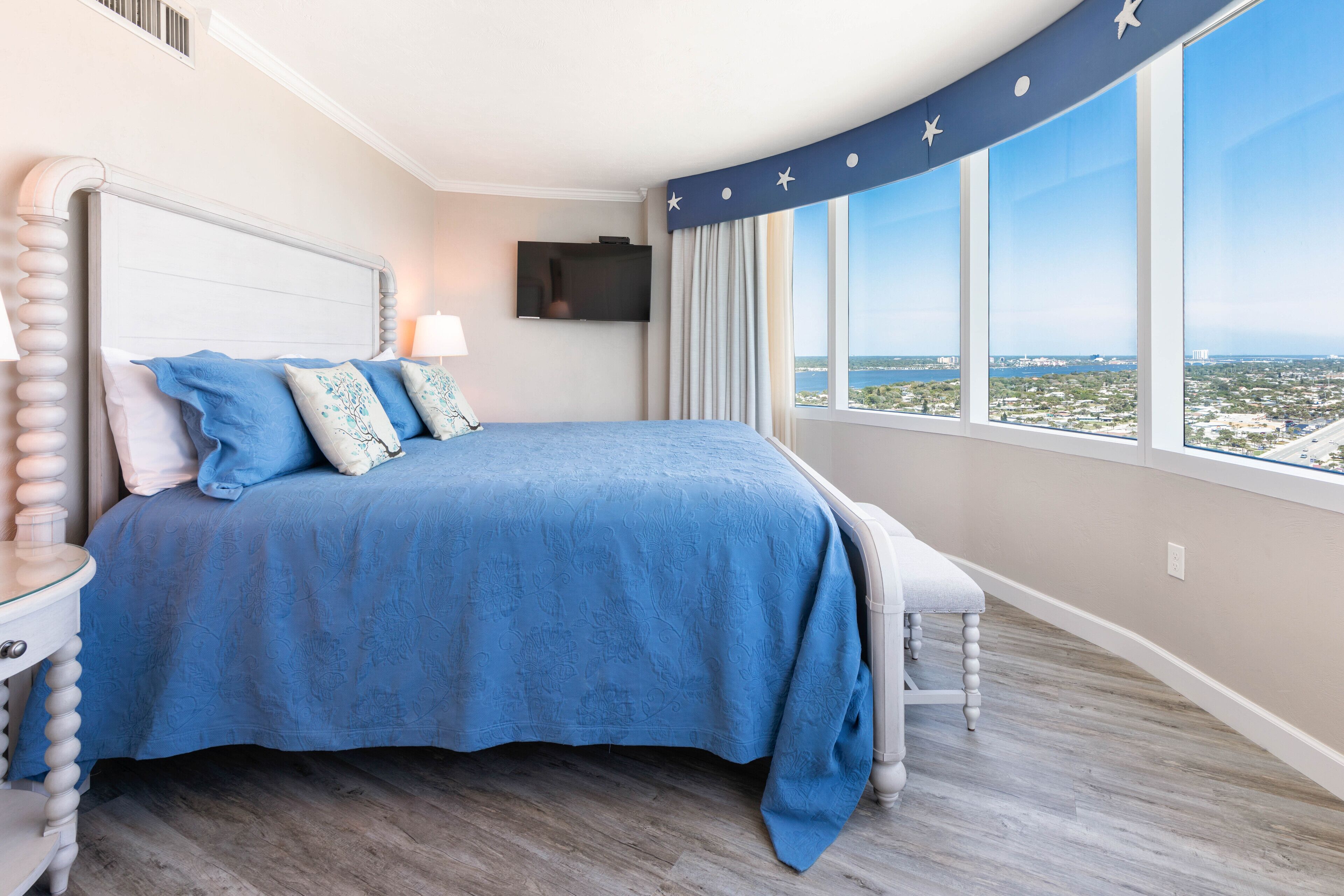 A bedroom with a large bed, a nightstand, a TV, and a window with a city view.